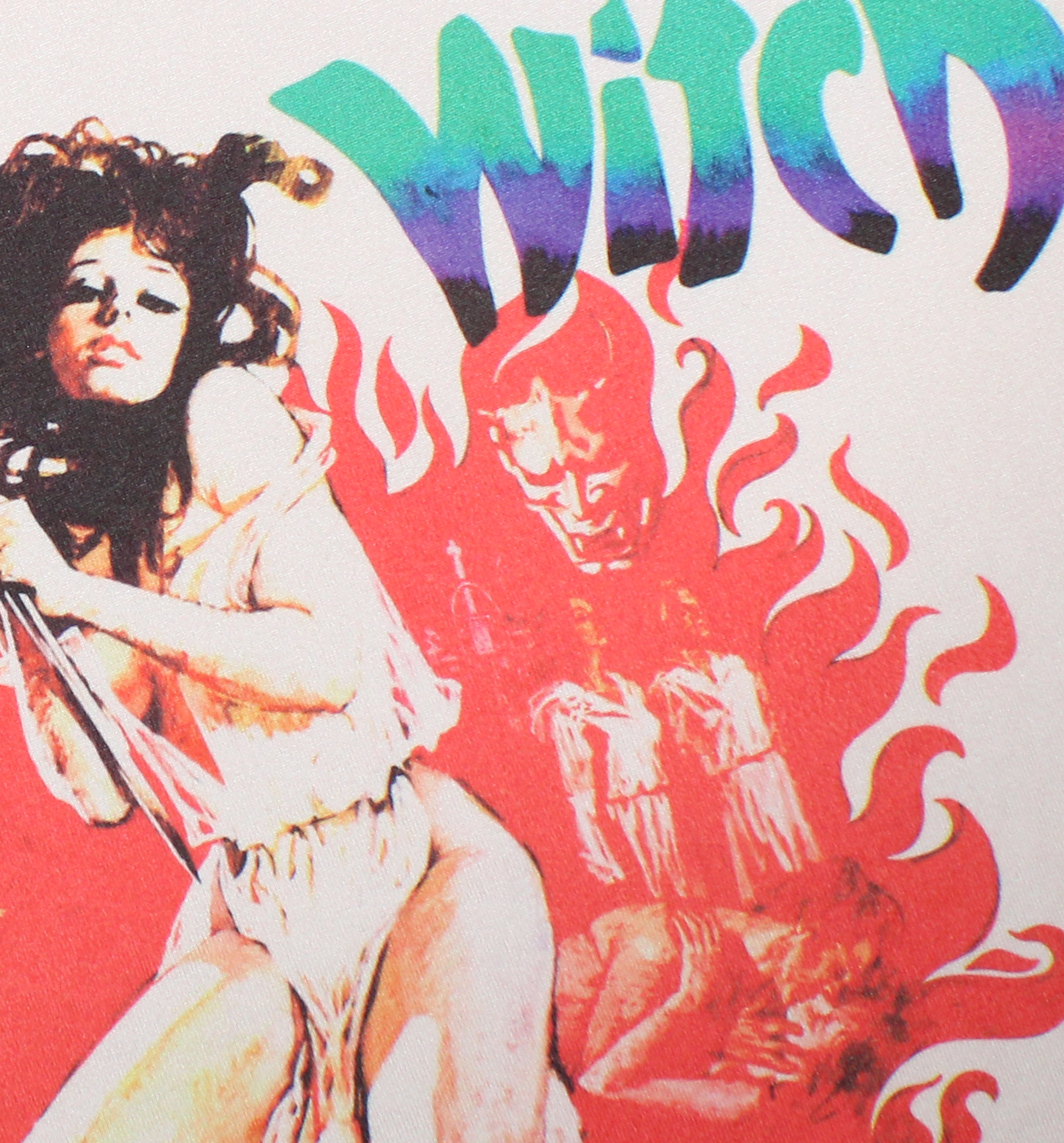A close-up of a graphic print featuring a stylized nude woman on the left, red flames, a demonic face in the center, and the word 'Witch' in large green-to-purple gradient lettering at the top. A woman wearing a beige ringer T-shirt with black trim and a colorful graphic print featuring the text 'Virgin Witch' and a red flame design witch a woman holding a knife. Virgin Witch horror tshirt. 70s horror shirts. women's horror tees tshirts. women's horror movie ringer shirt. witch ringer tshirt. occult horror 