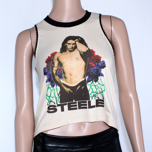 Women's Peter Steele relaxed fit, slouchy tank top. Type O Negative Top is tan/beige color, Asymmetrical hem, black lace-edged arms and black crew neck with an image of Peter Steele's Playgirl Cover. Band shirt.  Killer Kitty Clothing Alternative Brand. Mannequin wearing a sleeveless top with a graphic design and 'STEELE' text on a tan background