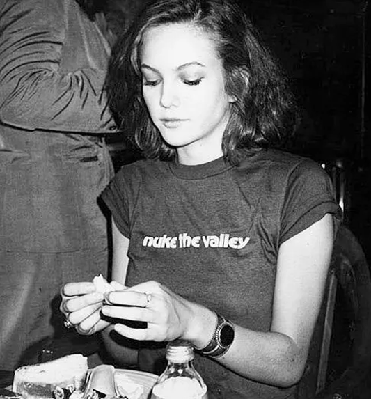 A black and white photograph of Diane Lane, photographed by Andy Warhol, wearing a dark graphic t-shirt with the text 'nuke the valley' across the chest, seated and holding food in a casual setting.