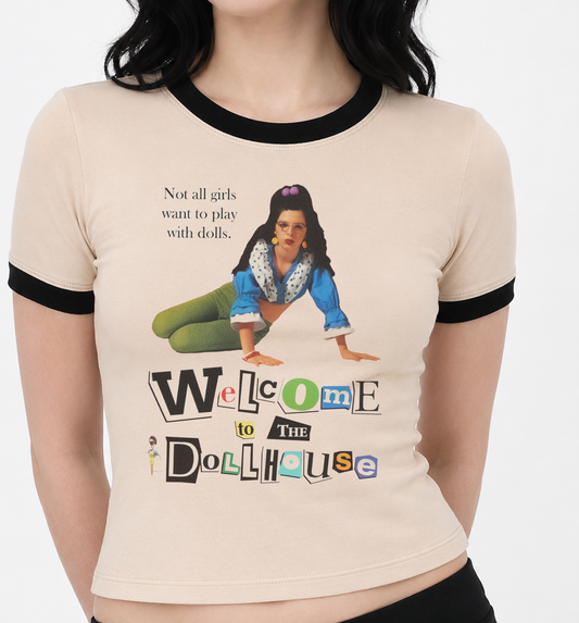A woman wearing a ringer t-shirt with a beige body, black collar and sleeve trim, featuring artwork from the 1995 cult classic  film Welcome to the Dollhouse 'Not all girls want to play with dolls' and 'Welcome to the Dollhouse'.

