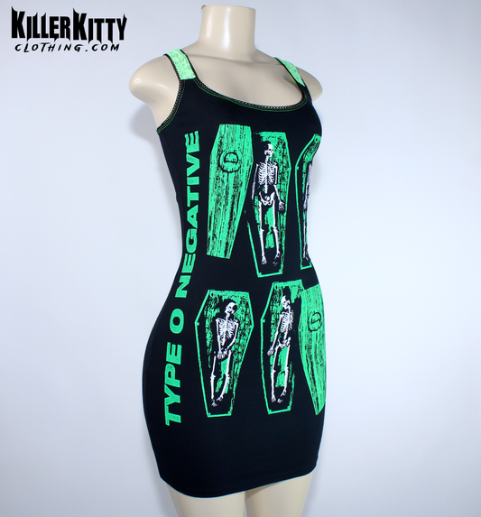 A sleeveless black mini dress with bright green trim and a graphic print of skeleton figures inside coffin shapes, displayed on a  mannequin. Type O negative dress for women. goth metal bodycon dress. peter steele goth metal dress. women's type o negative shirt. upcycled type o negative dress. heavy metal type o negative bodycon dress. diy alternative type o negative dress. killer kitty clothing upcycled band dresses. goth horror skeleton dress for women. alt fashion brand killer kitty. band dress for women