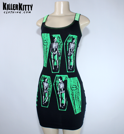 A sleeveless black mini dress with bright green trim and a graphic print of skeleton figures inside coffin shapes, displayed on a  mannequin. Type O negative dress for women. goth metal bodycon dress. peter steele goth metal dress. women's type o negative shirt. upcycled type o negative dress. heavy metal type o negative bodycon dress. diy alternative type o negative dress. killer kitty clothing upcycled band dresses. goth horror skeleton dress for women. alt fashion brand killer kitty. band dress for women